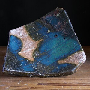 May include: A handcrafted ceramic plate with an unusual shape and glaze. The plate displays a mix of blue, green, and brown colours, with a textured surface and visible flaws. The design includes diagonal lines and speckled details, creating a rustic, artistic aesthetic.
