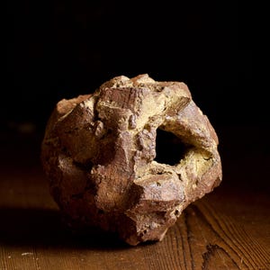 Könnte beinhalten: Eine strukturierte, braune Keramikskulptur mit einer großen, unregelmäßigen Öffnung. Das Stück hat ein raues, erdiges Aussehen, mit gelblichen Farbtönen und einem dunklen Hintergrund. Die Skulptur steht auf einer Holzoberfläche.
