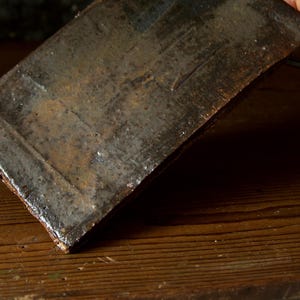 Puede incluir: Una placa rectangular de cerámica hecha a mano con una superficie oscura y texturizada. La placa tiene un aspecto rústico y envejecido con tonos marrones, grises y negros. Los bordes son ligeramente irregulares, lo que añade a su carácter único.