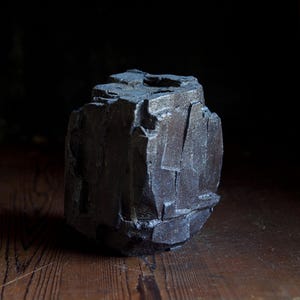 May include: A dark gray, roughly textured, geometric sculpture with a faceted surface. The object has a blocky, asymmetrical shape, and sits on a wooden surface. The sculpture's color is a deep charcoal.