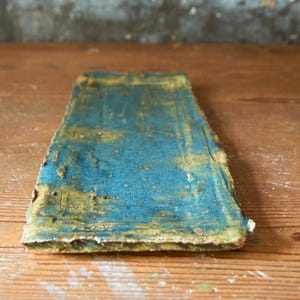 Puede incluir: Una pieza rectangular de cerámica hecha a mano con una superficie texturizada. La parte superior es de color azul verdoso moteado, con toques de dorado y marrón. Los bordes son ásperos e irregulares, lo que le da un aspecto rústico. Se muestra sobre una superficie de madera.