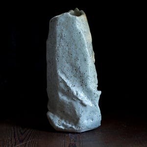 Könnte beinhalten: Eine hohe, strukturierte Keramikvase in Hellgrau mit gesprenkelten Details. Die Vase hat eine unregelmäßige, organische Form mit leicht ausgestellter Oberseite und rauer, unebener Oberfläche. Sie steht auf einer Holzoberfläche vor dunklem Hintergrund.