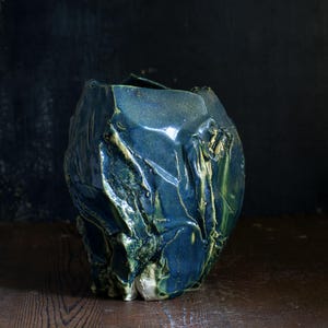 Könnte beinhalten: Eine handgefertigte, strukturierte Keramikvase mit einer einzigartigen, organischen Form. Die Vase hat eine tiefblaue Glasur mit grünen und goldenen Akzenten, die einen auffälligen Kontrast erzeugt. Die raue, unebene Oberfläche verstärkt ihren künstlerischen Reiz. Ein dekoratives Element für das Zuhause.