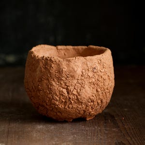 May include: A handmade, textured terracotta bowl. The bowl has a rounded shape with a rough, uneven surface. The colour is a warm reddish-brown. The bowl is sitting on a wooden surface.