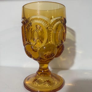Op de afbeelding: Een vintage amberkleurige glazen kelk. De basis heeft een sterrenpatroon, terwijl de kom een herhalend patroon van cirkels en sterren heeft. De steel is kort en het glas heeft een warme, gouden tint.