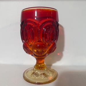 Pode incluir: Um cálice de vidro vermelho rubi vintage com um design de estrela na base. O cálice apresenta um padrão detalhado de círculos e estrelas, com uma haste e base em uma cor âmbar mais clara. O vidro tem um acabamento brilhante.