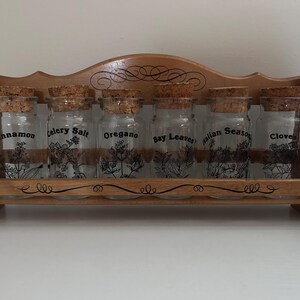 Puede incluir: Un especiero de madera con seis frascos de vidrio, cada uno etiquetado con el nombre de una especia en texto negro. Los frascos tienen tapones de corcho y están llenos de varias especias. El estante es de madera marrón claro.