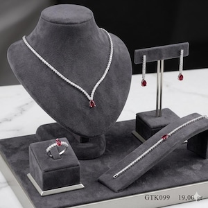 Peut inclure: Un ensemble de bijoux présenté sur une surface en velours gris. L'ensemble comprend un collier, des boucles d'oreilles, une bague et un bracelet, tous ornés de pierres transparentes et de pierres précieuses rouges en forme de goutte. Le collier est drapé sur un buste.