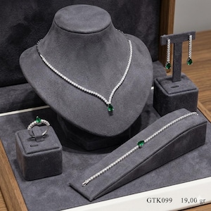 May include: A silver-toned jewellery set comprising a necklace, earrings, ring, and bracelet. Each piece is embellished with clear stones and emerald-green teardrop gems. The set is presented on a grey velvet display.