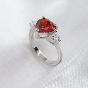 Może przedstawiać: Srebrny pierścionek z dużym, czerwonym, sercowatym kamieniem szlachetnym i dwoma mniejszymi, przezroczystymi kamieniami.