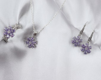 May include: A jewelry set featuring a ring, necklace, and earrings. Each piece has a silver-colored metal setting and is adorned with purple flower-shaped gemstones. The set is displayed on a white, textured fabric.