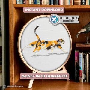 May include: A cross-stitch design of a calico cat, framed in a wooden embroidery hoop. The cat features orange, black, and white colours. The hoop is displayed on a wooden stand. The image includes the text "Instant Download", "Pattern Keeper Compatible", and "Money Back Guarantee".