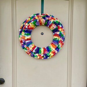 May include: A colorful fabric wreath with a rainbow of colors, including red, orange, yellow, green, blue, and purple. The wreath is hanging on a white door.