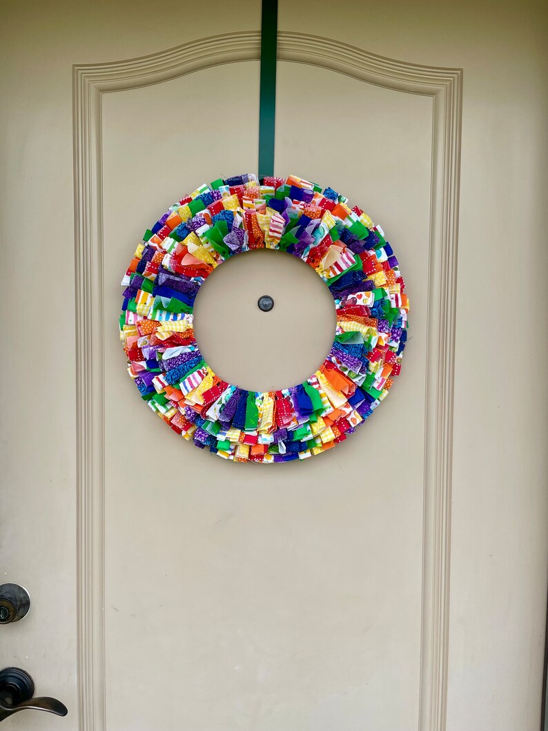 May include: A colorful fabric wreath with a rainbow of colors, including red, orange, yellow, green, blue, and purple. The wreath is hanging on a white door.