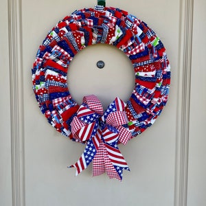 May include: A red, white, and blue fabric wreath with a matching bow. The wreath is made of strips of fabric sewn together in a circle. The bow is tied with a red, white, and blue gingham ribbon.