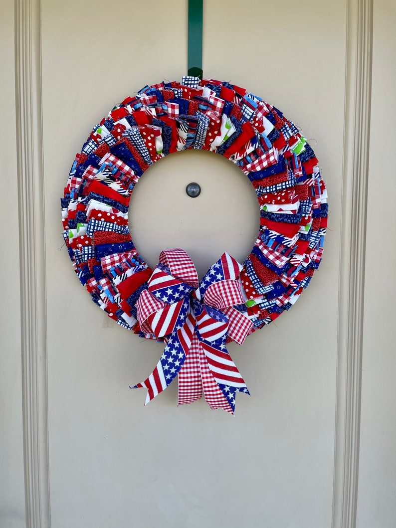 May include: A red, white, and blue fabric wreath with a matching bow. The wreath is made of strips of fabric sewn together in a circle. The bow is tied with a red, white, and blue gingham ribbon.