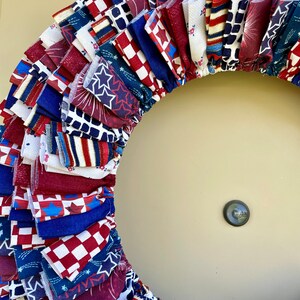 May include: A red, white, and blue fabric wreath with a variety of patriotic patterns, including stars, stripes, and fireworks. The wreath is made of multiple layers of fabric, creating a full and fluffy look.