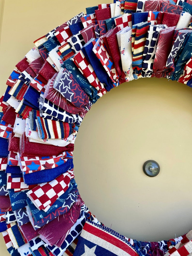 May include: A red, white, and blue fabric wreath with a variety of patriotic patterns, including stars, stripes, and fireworks. The wreath is made of multiple layers of fabric, creating a full and fluffy look.