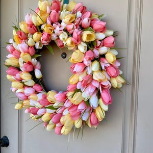 May include: A spring wreath made of pink, yellow and white artificial tulips. The wreath is made on a brown base and is decorated with green leaves.