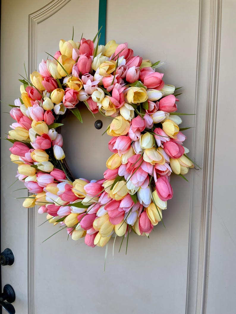 May include: A spring wreath made of pink, yellow and white artificial tulips. The wreath is made on a brown base and is decorated with green leaves.