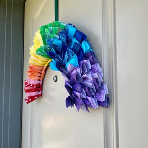 May include: A rainbow-colored fabric wreath with a red and white checkered bow hanging on a white door.
