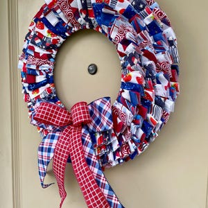 May include: A red, white, and blue fabric wreath with a red and blue plaid bow. The wreath is made of fabric scraps and is decorated with stars, stripes, and strawberries.