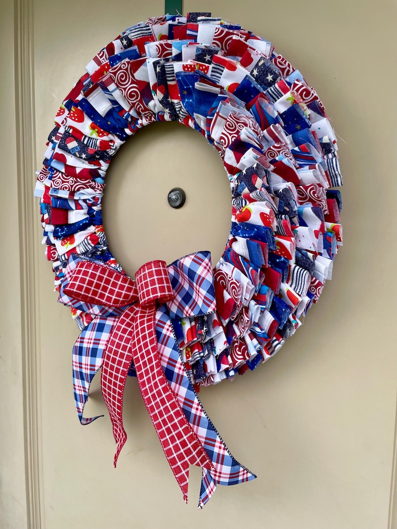 May include: A red, white, and blue fabric wreath with a red and blue plaid bow. The wreath is made of fabric scraps and is decorated with stars, stripes, and strawberries.
