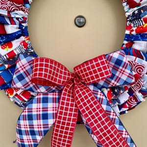 May include: A red, white, and blue fabric wreath with a red and blue plaid bow. The wreath is made of fabric scraps and is decorated with stars and stripes.