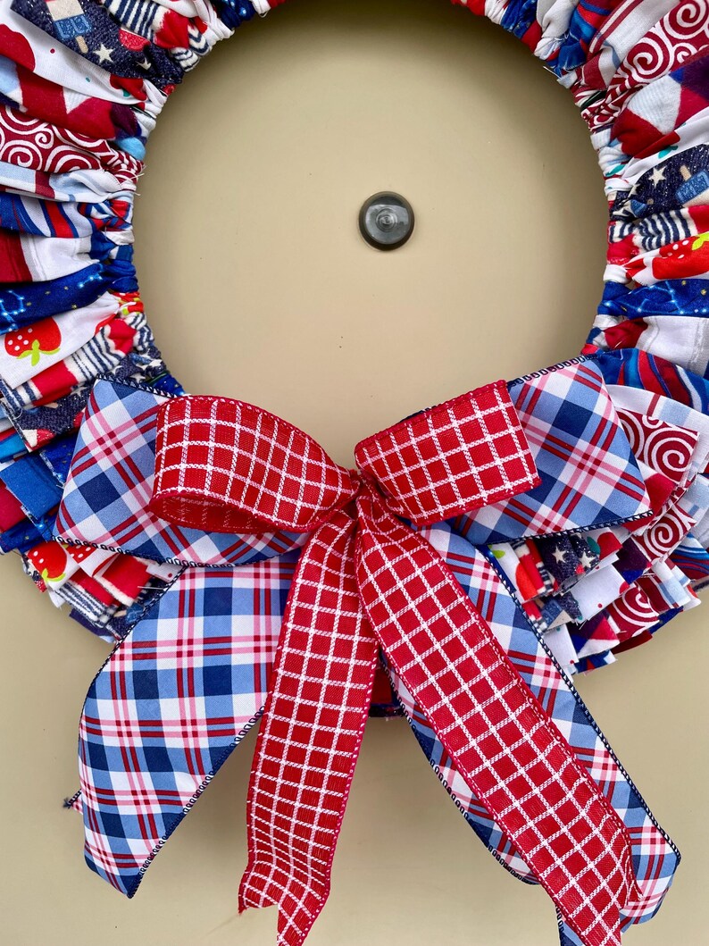 May include: A red, white, and blue fabric wreath with a red and blue plaid bow. The wreath is made of fabric scraps and is decorated with stars and stripes.