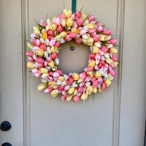 May include: A pink, yellow and white tulip wreath hanging on a light brown door. The wreath is made of artificial tulips and is a spring-themed decoration.