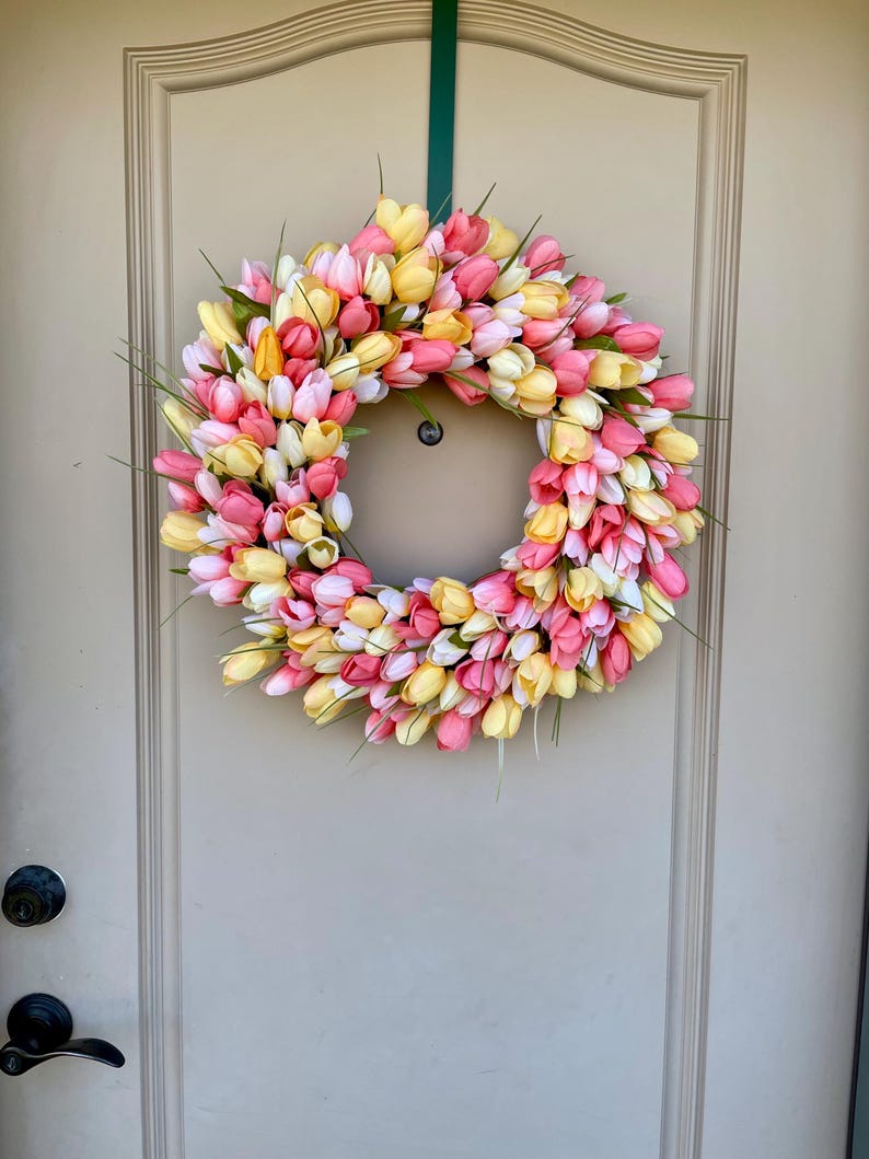 May include: A pink, yellow and white tulip wreath hanging on a light brown door. The wreath is made of artificial tulips and is a spring-themed decoration.