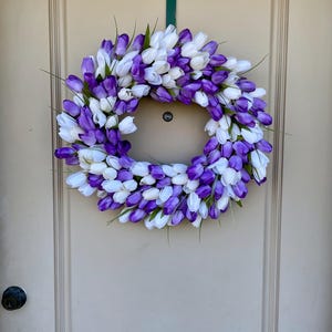May include: A purple and white tulip wreath hanging on a light beige door. The wreath is made of artificial tulips and is a great way to add a touch of spring to your home.