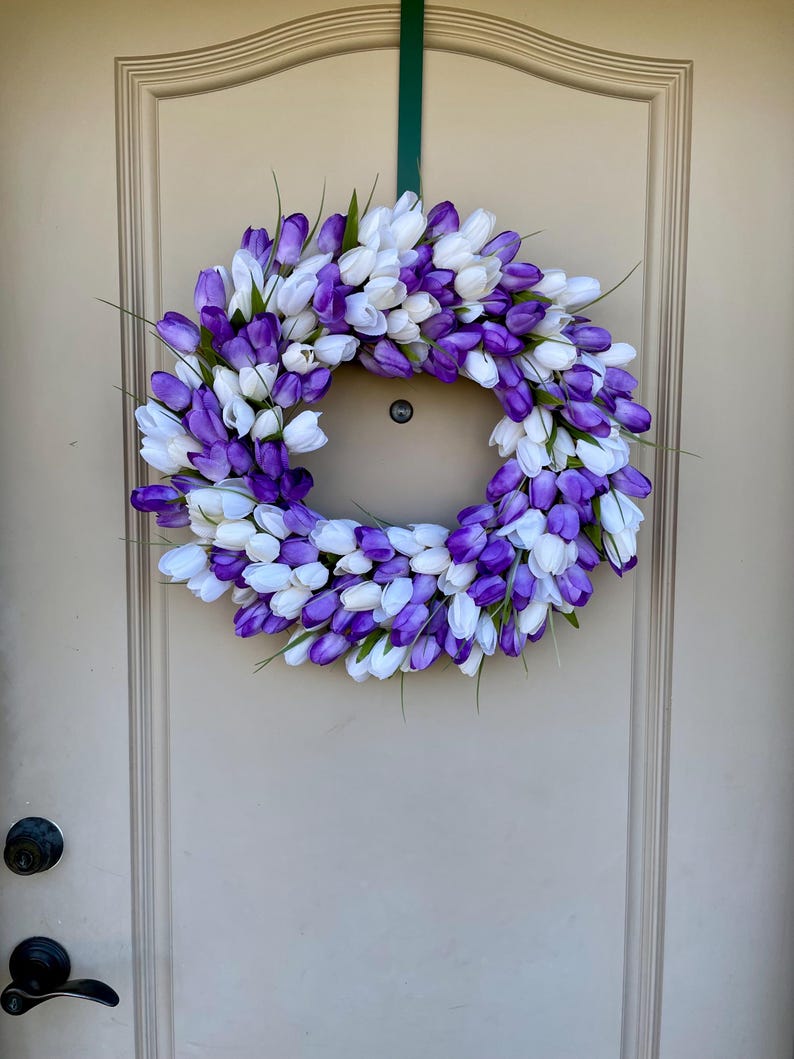 May include: A purple and white tulip wreath hanging on a light beige door. The wreath is made of artificial tulips and is a great way to add a touch of spring to your home.