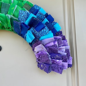 May include: A rainbow-colored fabric wreath with green, blue, purple, and gold fabric scraps. The wreath is made of fabric scraps sewn together in a ruffled style.