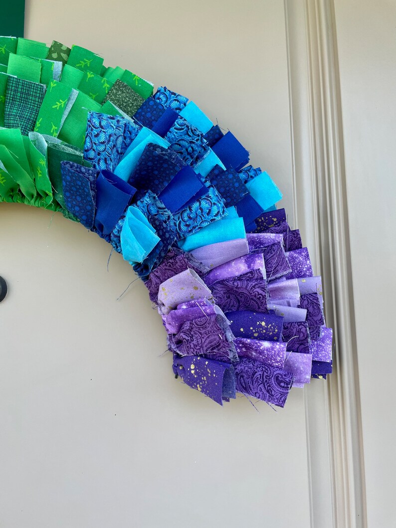 May include: A rainbow-colored fabric wreath with green, blue, purple, and gold fabric scraps. The wreath is made of fabric scraps sewn together in a ruffled style.