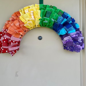 May include: A rainbow-colored fabric wreath with red, orange, yellow, green, blue, purple, and white fabric pieces. The wreath is hanging on a white door.