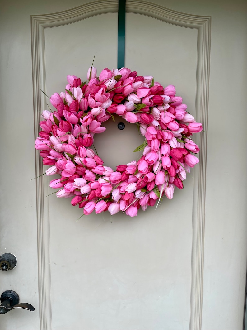 May include: A pink and white tulip wreath hanging on a white door. The wreath is made of artificial tulips and is a great way to add a touch of spring to your home.