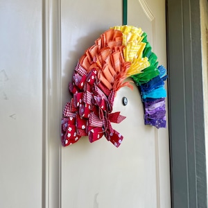 May include: A rainbow-colored fabric wreath with red, orange, yellow, green, blue, and purple fabric strips. The wreath is hanging on a white door.