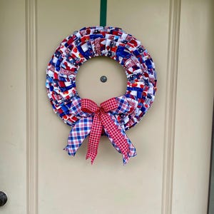 May include: A red, white, and blue fabric wreath with a red and white plaid bow. The wreath is made of fabric scraps and is decorated with stars and stripes.
