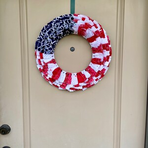 May include: A red, white, and blue fabric wreath with a ruffled design. The wreath is hanging on a white door.