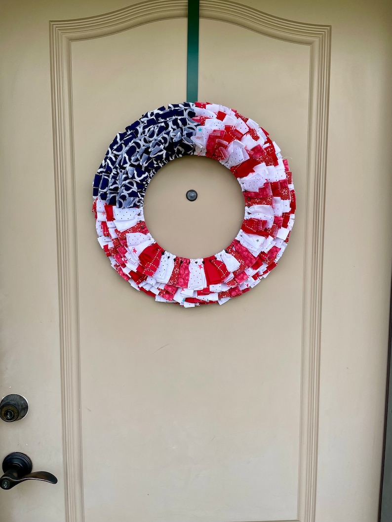 May include: A red, white, and blue fabric wreath with a ruffled design. The wreath is hanging on a white door.