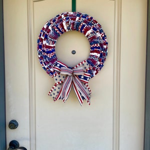 May include: A red, white, and blue fabric wreath with a star-patterned bow. The wreath is hanging on a white door.