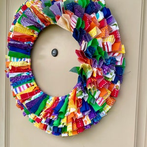 May include: A colorful fabric wreath with a rainbow of colors, including red, orange, yellow, green, blue, indigo, and violet. The wreath is made of many small fabric strips that are tied together to create a ruffled effect.