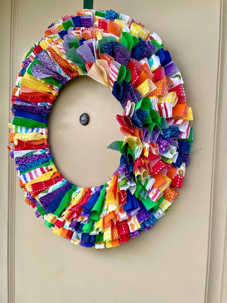 May include: A colorful fabric wreath with a rainbow of colors, including red, orange, yellow, green, blue, indigo, and violet. The wreath is made of many small fabric strips that are tied together to create a ruffled effect.