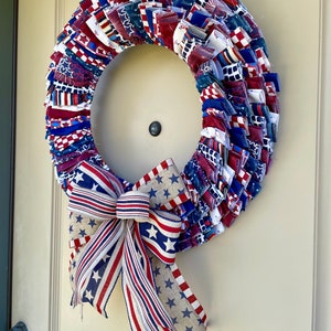 May include: A red, white, and blue fabric wreath with a matching bow. The wreath is made of strips of fabric with stars and stripes patterns. The bow is tied with a burlap ribbon.