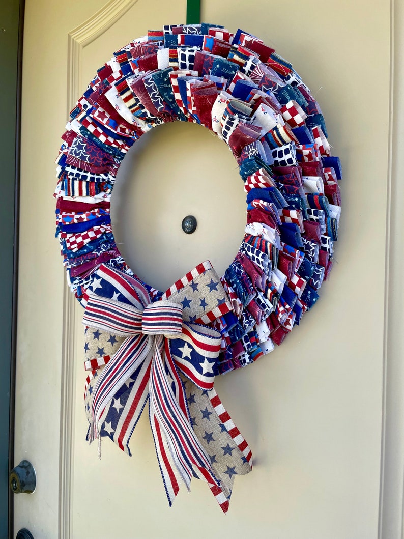 May include: A red, white, and blue fabric wreath with a matching bow. The wreath is made of strips of fabric with stars and stripes patterns. The bow is tied with a burlap ribbon.