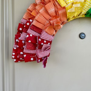 May include: A colorful fabric wreath with red, white, orange, yellow, and green fabric strips. The wreath is made of multiple layers of fabric, creating a ruffled effect.