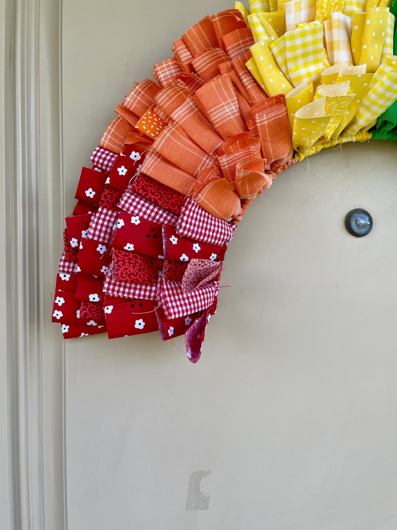 May include: A colorful fabric wreath with red, white, orange, yellow, and green fabric strips. The wreath is made of multiple layers of fabric, creating a ruffled effect.