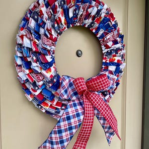 May include: A red, white, and blue fabric wreath with a plaid bow. The wreath is made of scraps of fabric sewn together in a ruffled style. The bow is made of red and blue plaid ribbon.