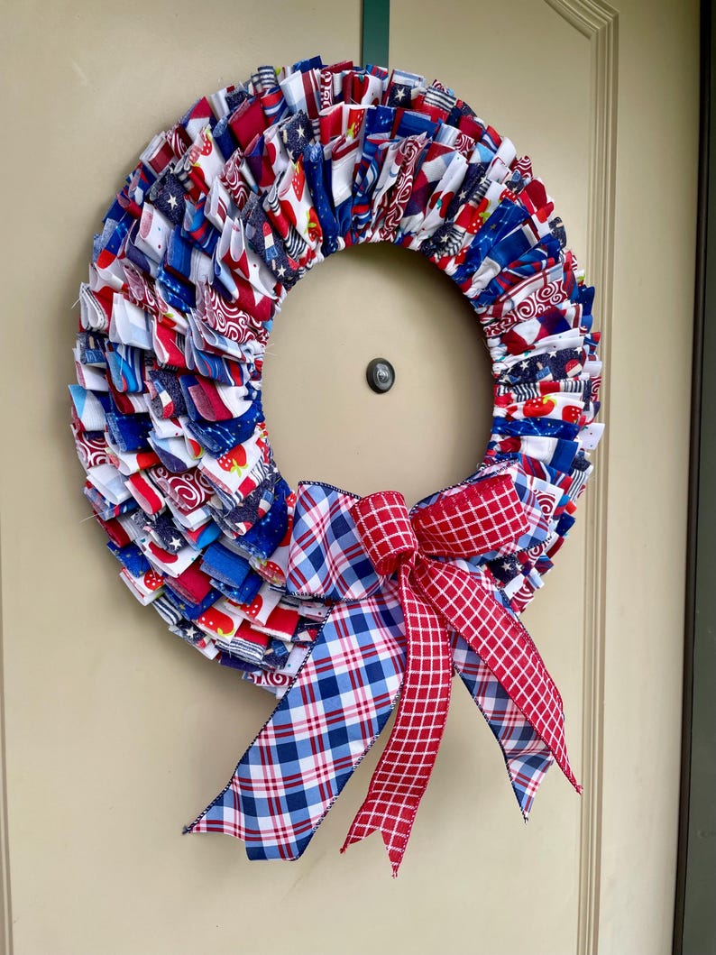 May include: A red, white, and blue fabric wreath with a plaid bow. The wreath is made of scraps of fabric sewn together in a ruffled style. The bow is made of red and blue plaid ribbon.